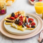 Two tender crepes made with an easy blend-and-pour method are folded and topped with berries and cream, served on a plate beside a jar of jam, a glass of orange juice, and a bowl of mixed berries.