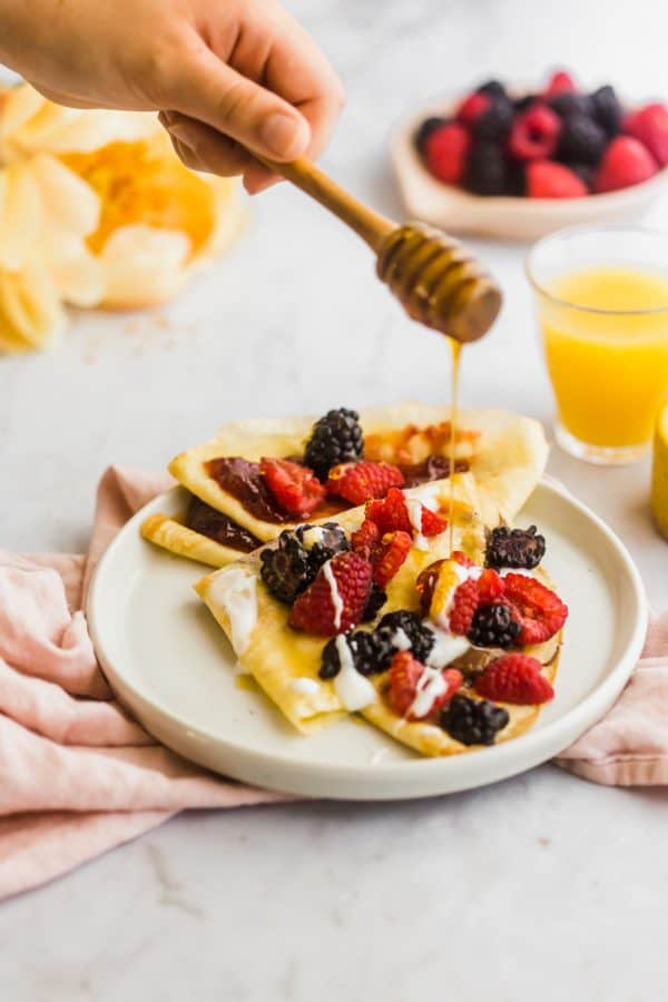 A hand drizzles honey over crepes made with an easy crepe recipe, topped with whipped cream and mixed berries on a white plate, with juice and fruit in the background.
