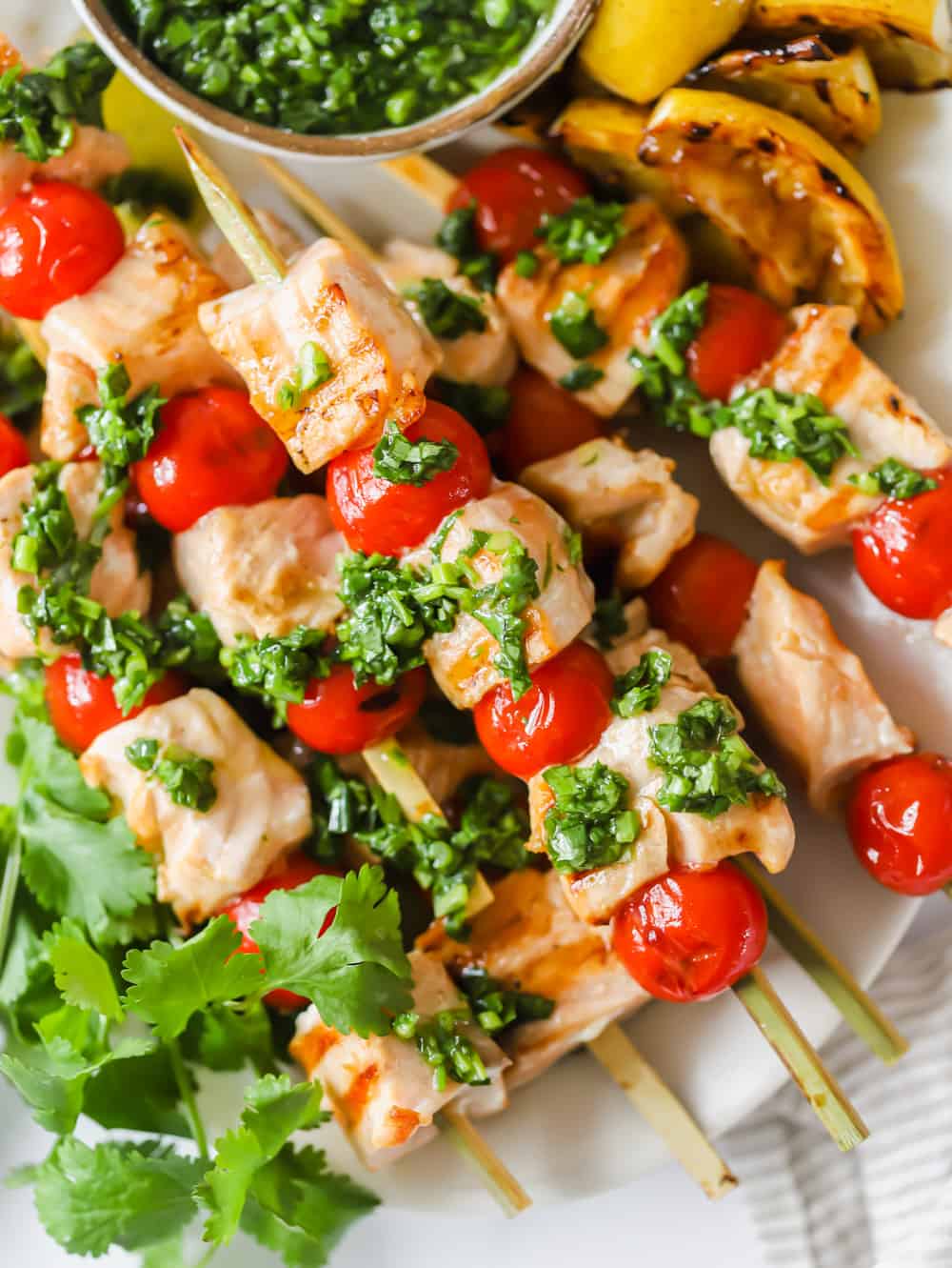 Enjoy these Grilled Salmon Kabobs for a quick and easy lunch or dinner. Juicy salmon and tomatoes are threaded onto skewers before being grilled to perfection and topped with chimichurri sauce. It’s a simple, yet flavor-packed meal! #salmonkabobs #summerrecipe #grilling #grilledsalmon #kabobs close up on salmon and tomato skewers topped with green herb sauce.