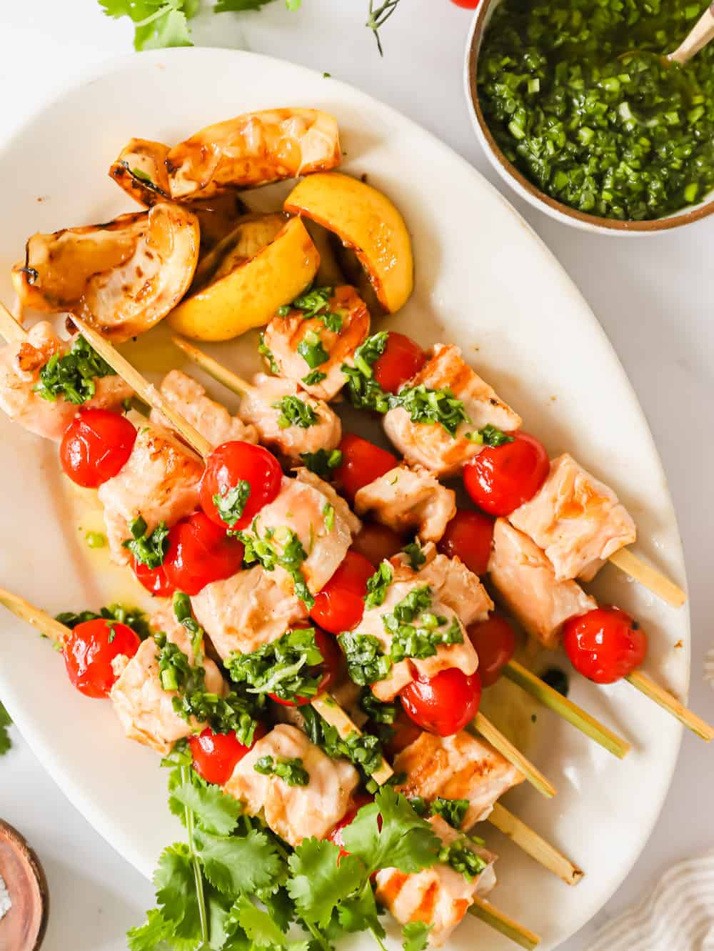 Enjoy these Grilled Salmon Kabobs for a quick and easy lunch or dinner. Juicy salmon and tomatoes are threaded onto skewers before being grilled to perfection and topped with chimichurri sauce. It’s a simple, yet flavor-packed meal! #salmonkabobs #summerrecipe #grilling #grilledsalmon #kabobs salmon and tomato skewers on a white serving plate next to a small bowl of green herb sauce.