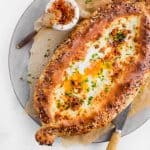 Oval-shaped Khachapuri, a cheesy bread filled with gooey egg and melted cheese, garnished with herbs, served on parchment paper with a knife and a small dish of spices.