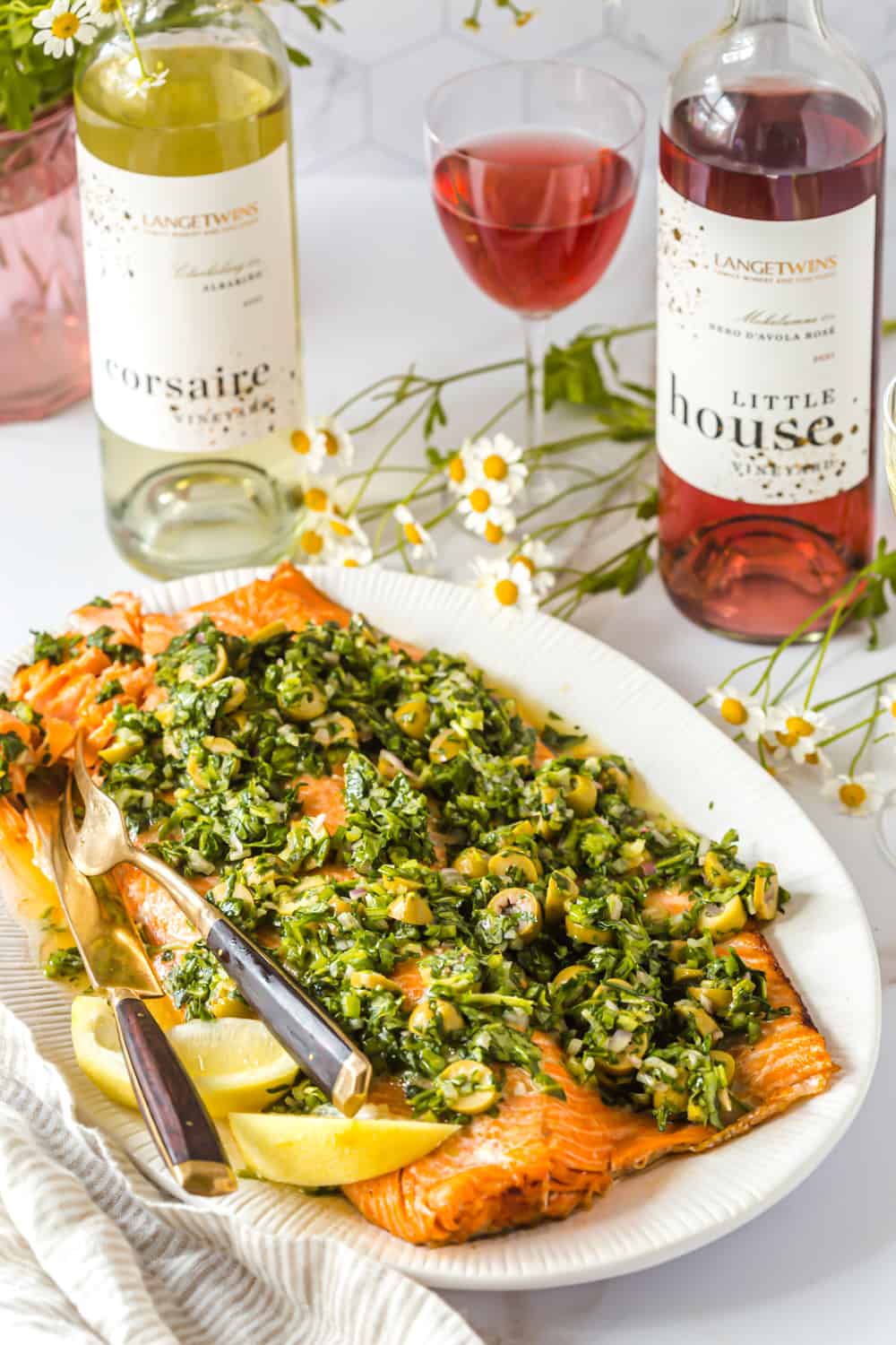 oven baked trout topped with herb olive salsa on a white serving platter next to 2 bottles of wine.