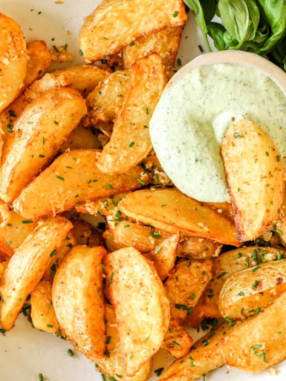 These Air Fryer Potato Wedges are tossed with savory seasonings and parmesan cheese, leaving you with an irresistibly crispy and crunchy snack or side dish. Take them to the next level with homemade basil aioli for dipping! #potatowedges #airfryer #easysnack #sidedish close up on potato wedges being dipped into a small bowl of basil aioli.