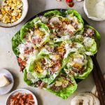 A plate of butter lettuce cups filled with grilled corn, sliced radishes, prosciutto bits, shredded cheese, and creamy dressing.