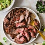 Sliced grilled frank steak carne asada on a white plate with a gold fork, surrounded by guacamole, chopped onions and cilantro, lime, and tortillas.