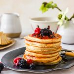 A stack of pancakes topped with mixed berries and syrup sits on a black plate with a fork, with a cup, jug, and flowers in the blurred background.