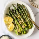 Roasted asparagus spears topped with grated cheese, minced garlic, and seasonings, served with a wedge of lemon on a white plate.