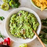 A bowl of simple guacamole with a spoon, surrounded by lime halves, cilantro, an avocado, tomatoes, and a bowl of tortilla chips.