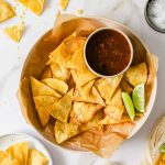 A plate of tortilla chips with a small bowl of salsa and lime wedges on parchment paper.