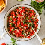 A bowl of freshly made pico de gallo with diced tomatoes, onions, cilantro, and jalapeño, surrounded by lime, salt, chips, and fresh ingredients.