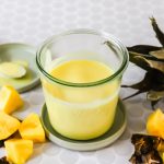 A glass jar filled with yellow pineapple dressing sits on a matching saucer, surrounded by pineapple chunks and pineapple leaves.