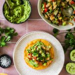 A tostada topped with mashed avocado and shrimp salad sits on a plate, surrounded by bowls of mashed avocado, shrimp salad, lime halves, cilantro, salt, and stacked tostadas on a pink tiled surface.