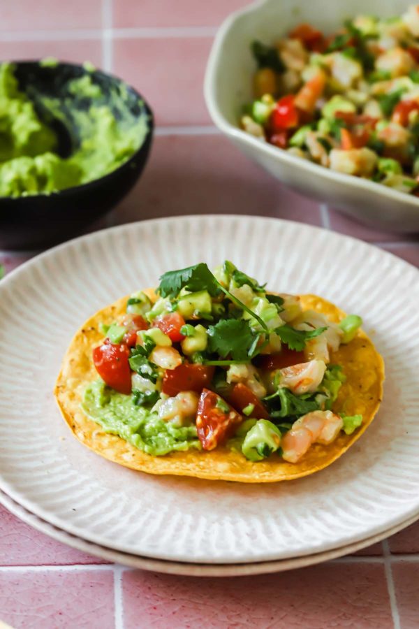 A tostada topped with guacamole, shrimp, diced tomatoes, avocado, and cilantro sits on a plate, with bowls of guacamole and shrimp mixture in the background.