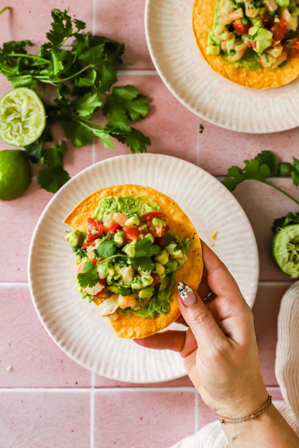 A hand holds a tostada topped with guacamole, diced tomatoes, and cilantro over a plate on a pink tiled surface with limes and cilantro in the background.