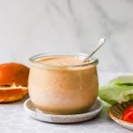 A glass jar of creamy, burger sauce with a spoon inside, placed on a small plate next to lettuce, tomato, and a hamburger buns.