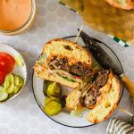 A crack burger cut in half on a plate with lettuce, tomato, sauce, and pickles, next to a knife and a bowl of sliced pickles and tomatoes.