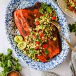 Grilled salmon fillet on a blue and white platter, topped with corn, tomato, and avocado salsa, garnished with cilantro and lime halves.
