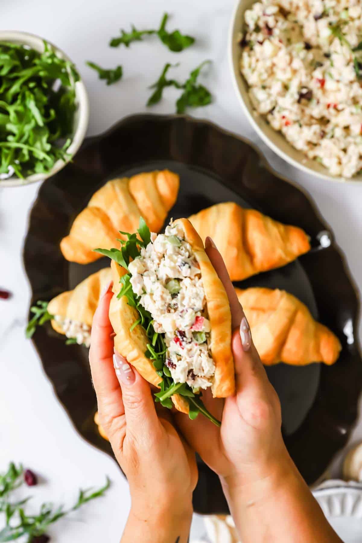 Hands holding a croissant sandwich filled with chicken salad and arugula; more croissant sandwiches, arugula, and a bowl of chicken salad are on the table.
