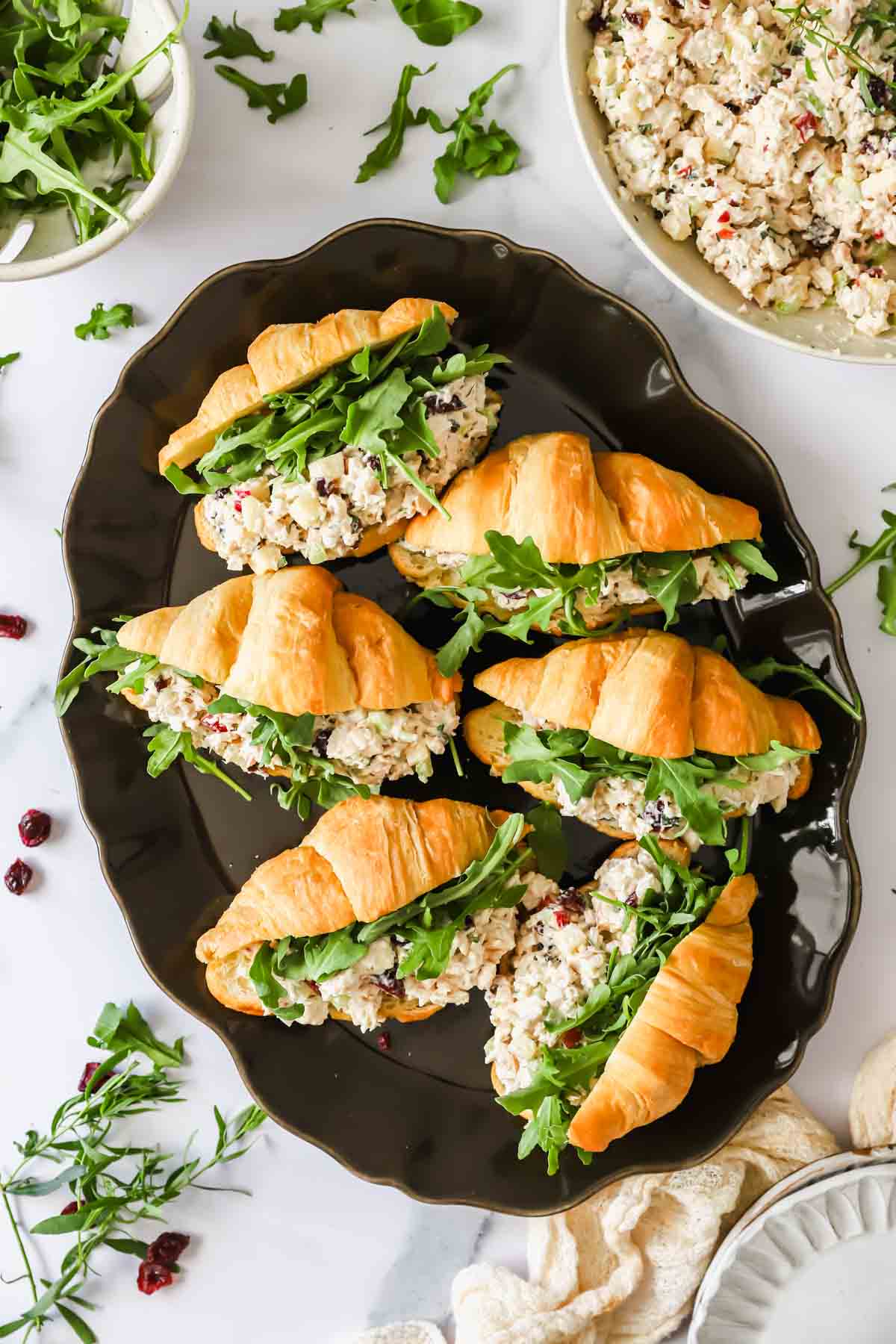 Five croissants filled with chicken salad and arugula are arranged on a dark platter, with extra greens and chicken salad in bowls nearby.
