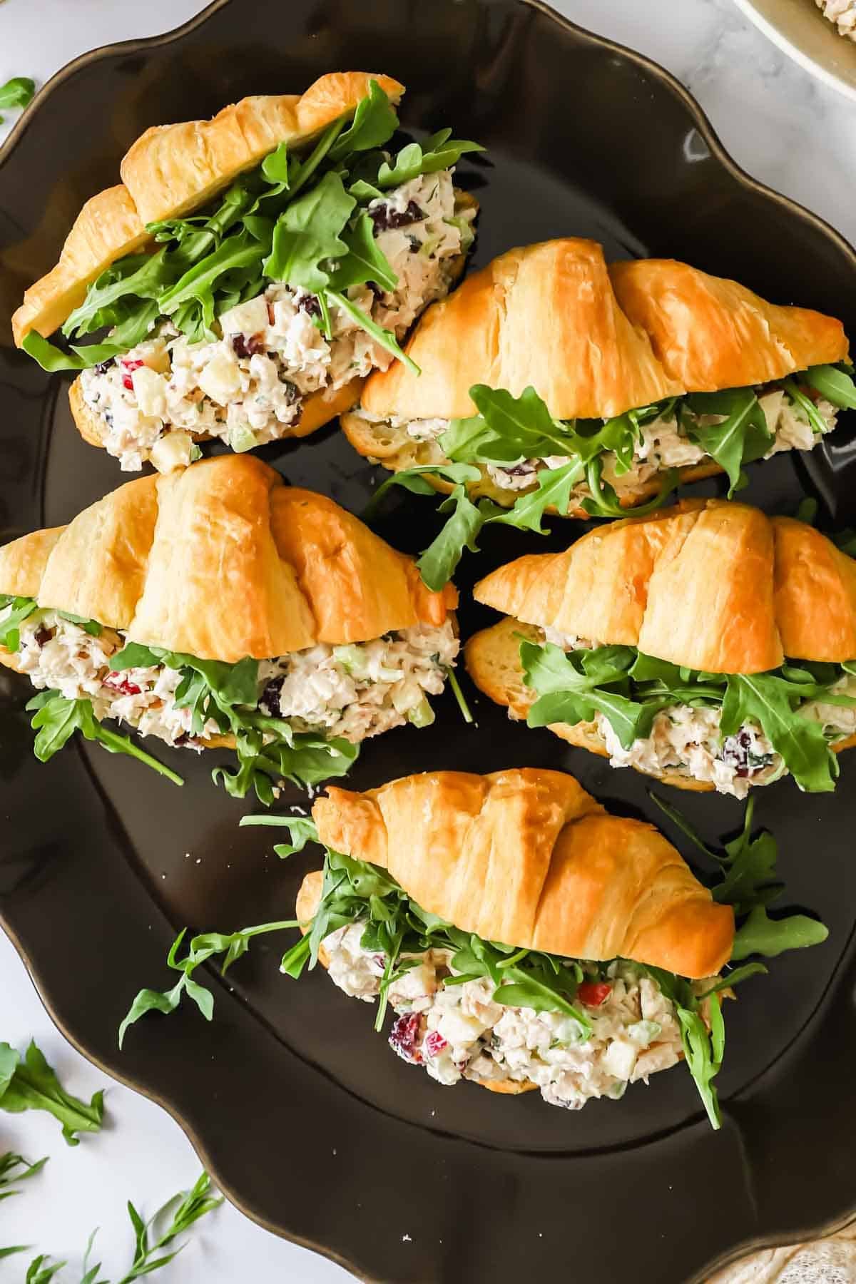 Five croissant sandwiches filled with chicken salad and fresh arugula are arranged on a dark plate.