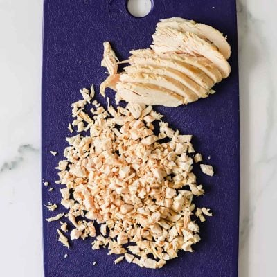 Sliced and finely chopped cooked chicken breast on a dark blue cutting board, placed on a light marble surface.