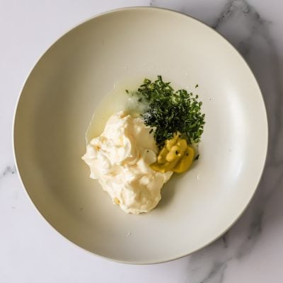 A white bowl containing mayonnaise, chopped fresh herbs, yellow mustard, and a small amount of lemon juice on a marble surface.