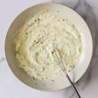 A white bowl containing a creamy, herb-flecked sauce with a small whisk resting inside, placed on a marble surface.