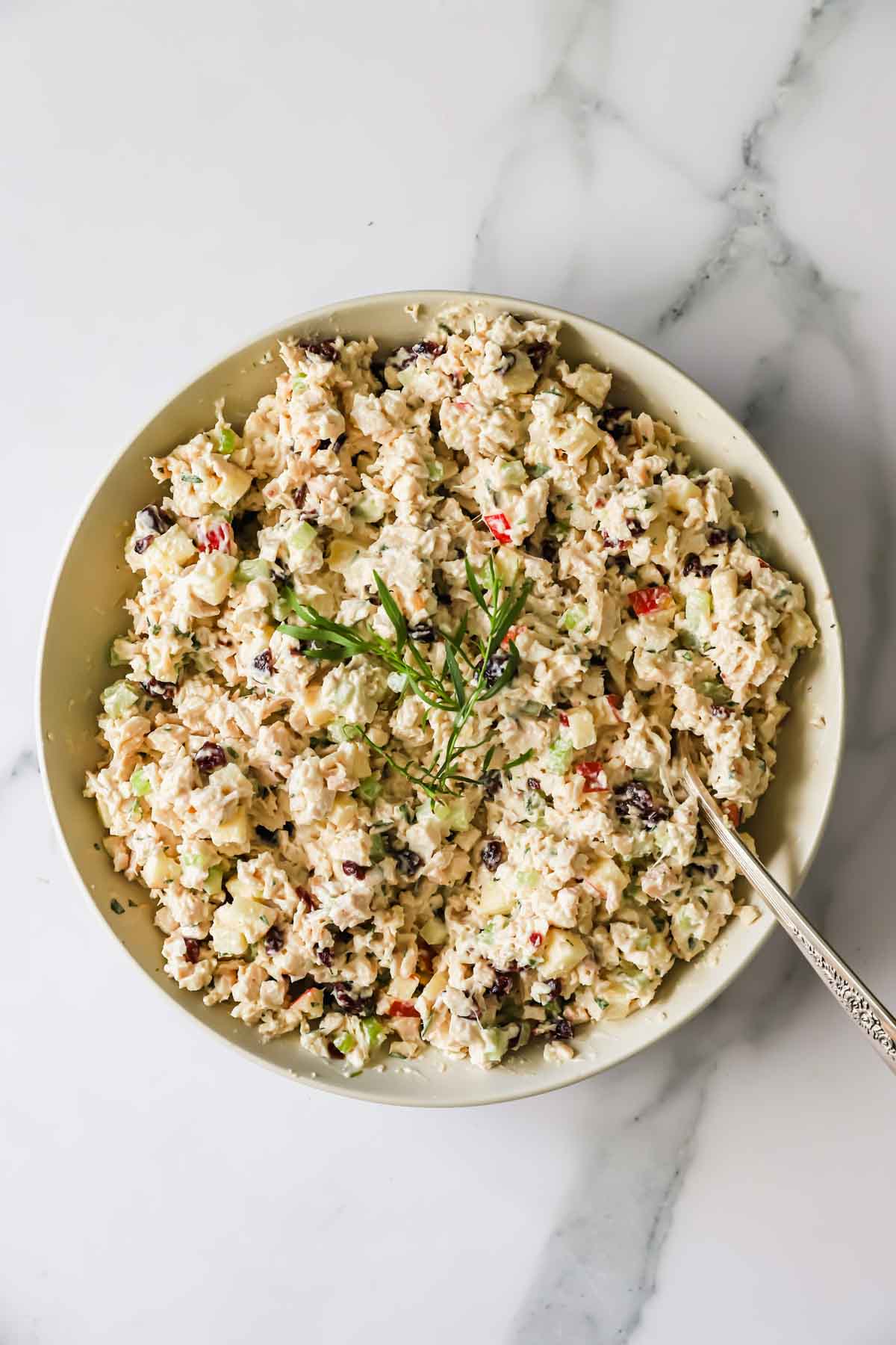 A bowl of chicken salad with chopped vegetables and dark bits, garnished with herbs, and a spoon resting inside, on a marble surface.