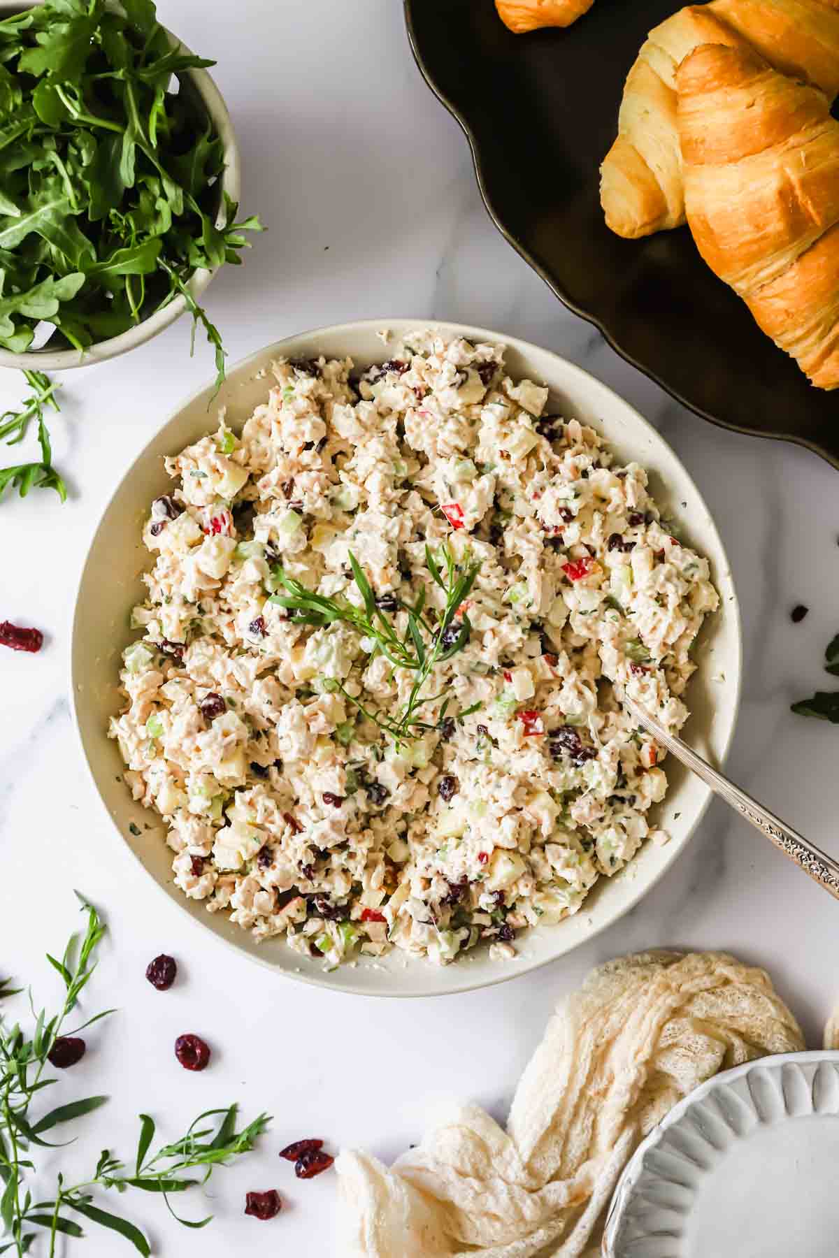 A bowl of chicken salad with herbs, chopped vegetables, and dried cranberries sits on a white surface next to croissants and a bowl of leafy greens.