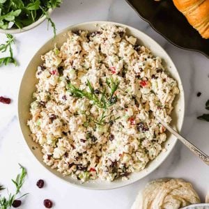 A bowl of chicken salad with chopped vegetables and herbs, garnished with fresh greens, sits on a marble surface with a spoon in the bowl.