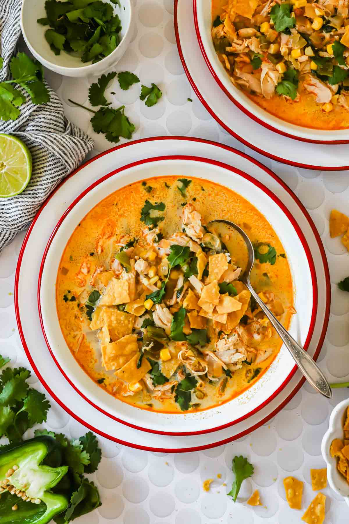 A bowl of creamy chicken poblano soup topped with tortilla strips, cilantro, and corn, with a spoon inside. Fresh cilantro, lime, and jalape&ntilde;o are on the table nearby.