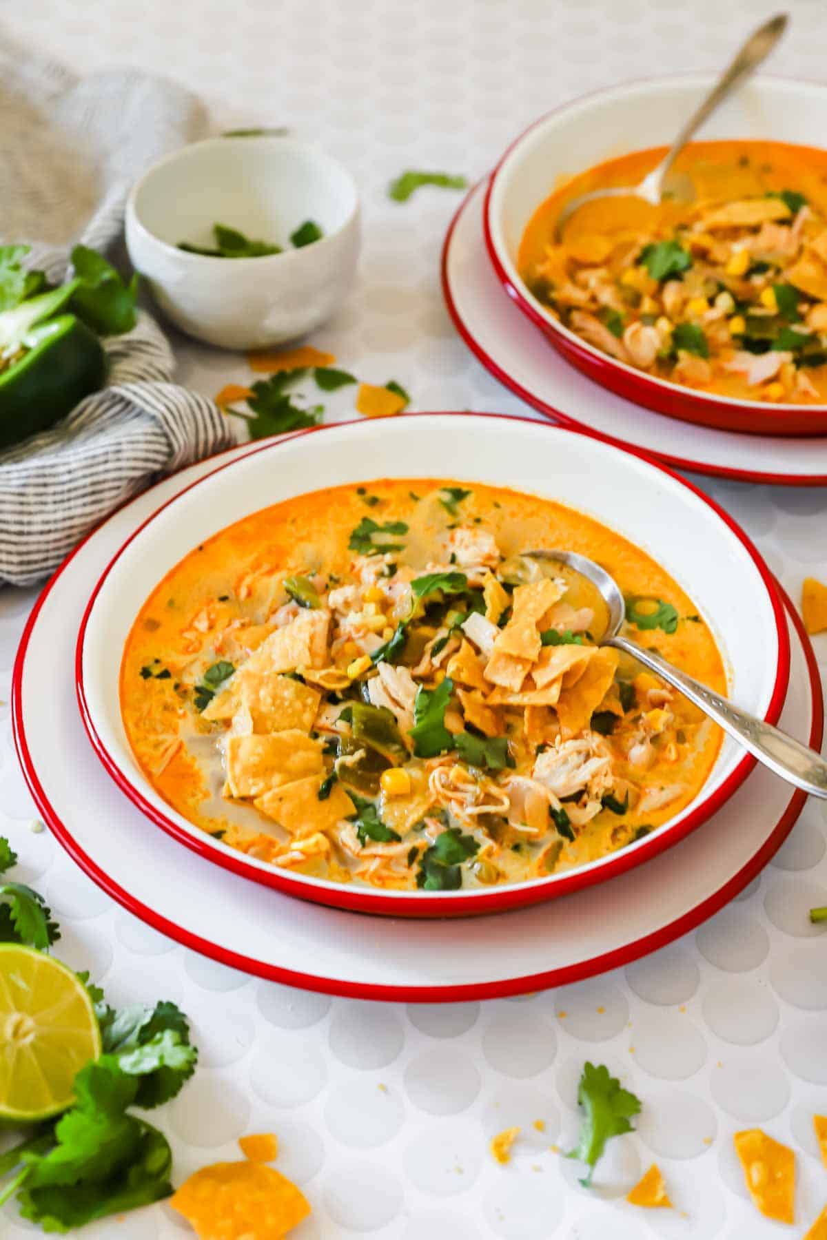Two bowls of creamy chicken poblano soup topped with tortilla chips and cilantro, with spoons in each bowl, set on a white table with lime wedges and herbs nearby.