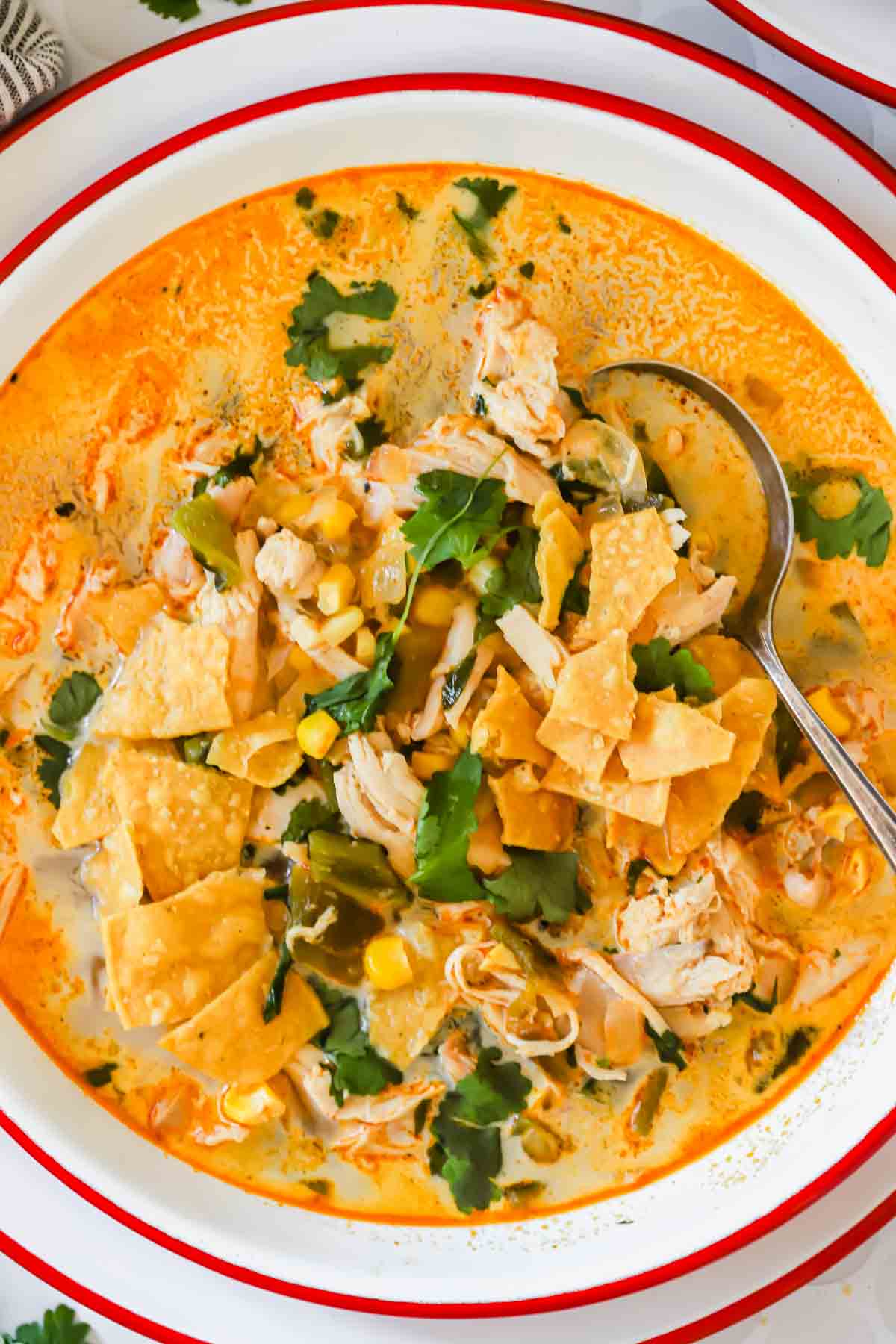 A bowl of chicken poblano soup with corn, tortilla chips, chopped cilantro, and a spoon, served on a white plate with a red rim.