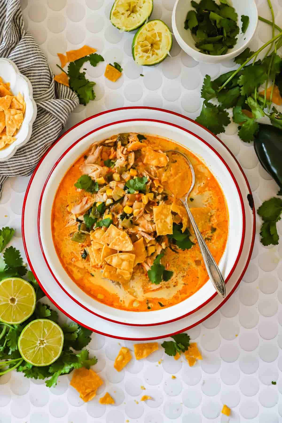 A bowl of creamy chicken poblano soup with shredded chicken, corn, herbs, and tortilla chips, garnished with cilantro. Lime halves and chopped cilantro are placed nearby on the table.