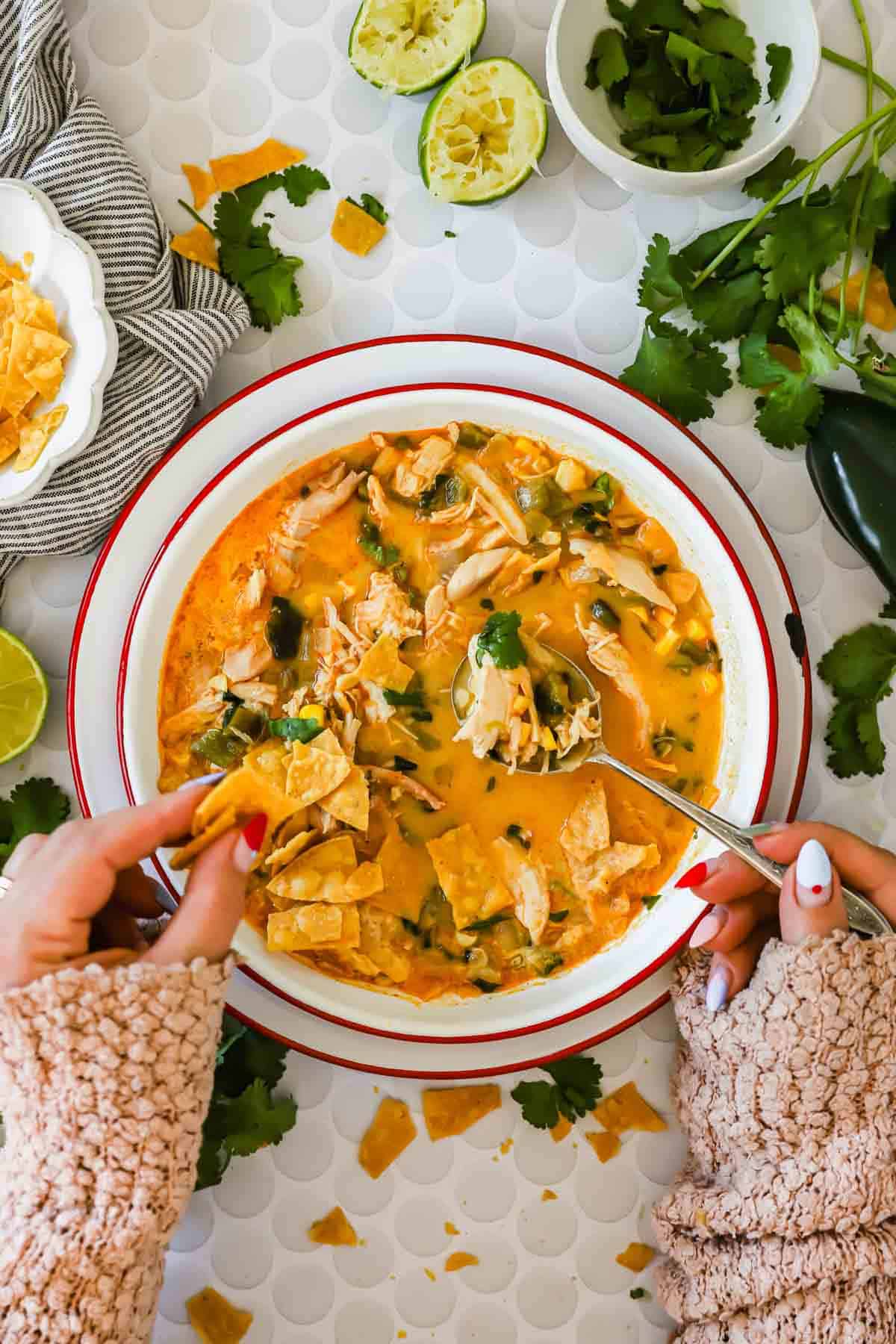 A bowl of creamy chicken poblano soup topped with cilantro and tortilla chips, surrounded by limes, cilantro, and a jalape&ntilde;o on a patterned surface. A person dips a chip into the soup.