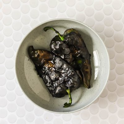 A gray bowl containing three charred poblano peppers sits on a white, textured surface with a circular pattern.