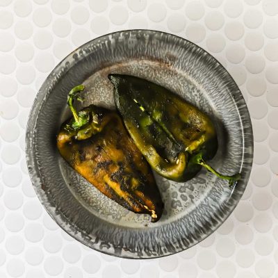 Two charred poblano peppers on a gray, round plate, placed on a white textured surface.