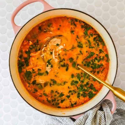 A pot of chicken poblano soup with shredded chicken and chopped herbs, placed on a white textured surface with a striped cloth nearby and a gold ladle inside.