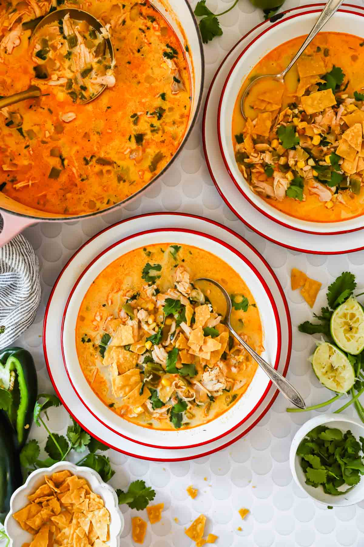 Two bowls and a pot of creamy chicken poblano soup garnished with cilantro and tortilla chips, surrounded by fresh lime, jalape&ntilde;o, and herbs on a white surface.