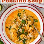 A bowl of chicken poblano soup topped with tortilla strips and cilantro, with a spoon, surrounded by fresh cilantro and lime wedges on a white table.