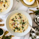 A bowl of creamy kielbasa potato soup with sliced sausage, potatoes, herbs, and dill garnish, with a spoon in the bowl, set on a white marble surface.