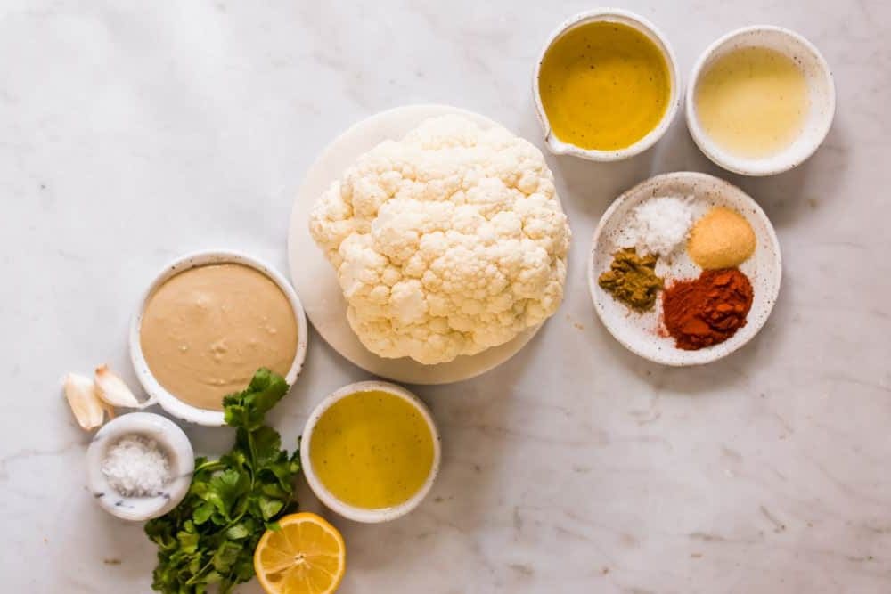 a head of cauliflower next to small white bowls of oil, sauce, herbs, and salt.