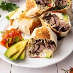 A plate with sliced steak burritos filled with rice, black beans, and avocado, served with sliced avocado and a side of pico de gallo.
