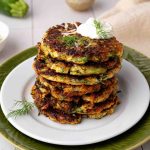 A stack of golden-brown zucchini fritters topped with a dollop of sour cream and a sprig of dill, served on a white plate with green garnish.
