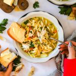 A person eating rotisserie chicken noodle soup with a spoon, holding buttered bread, with a striped napkin and herbs on a white table.
