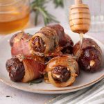 Bacon-wrapped dates stuffed with cheese on a plate, with honey being drizzled over them using a honey dipper.