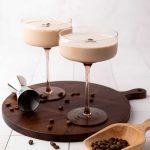 Two creamy espresso martinis in coupe glasses on a wooden board, with scattered coffee beans, a jigger, and a wooden scoop with more beans.