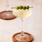A cocktail in a coupe glass garnished with four green olives on a skewer, placed on a wooden coaster.