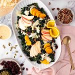 A white oval platter with fall harvest salad topped with apple slices, orange slices, dried cranberries, pecans, pumpkin seeds, and cheese shavings, surrounded by salad ingredients.