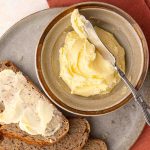 A bowl of butter with a butter knife on a plate, next to slices of whole grain bread, one slice partially spread with butter.